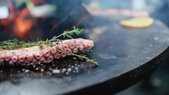 Octopus tentacle grilling on hot charcoal grill with lemon slices and fresh thyme. Seafood barbecue cooking over open fire flames. Outdoor summer food preparation concept.