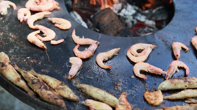 Shrimp and fish frying on hot griddle over open fire. Street food cooking process with flames and coals. Fresh seafood preparation at outdoor market grill.