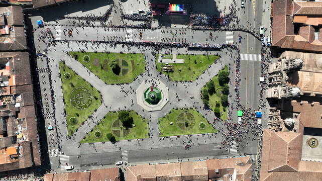 Aerial view of the historic Plaza de Armas in Cusco featuring colonial architecture, green gardens, stone walkways, and crowds of people surrounded by terracotta rooftops in Cusco, Peru.