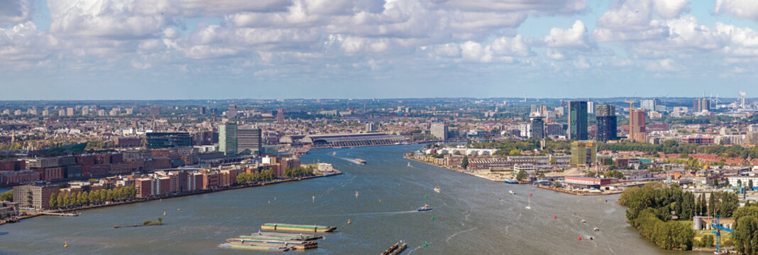 Aerial view of the IJ river flowing through Amsterdam with modern skyscrapers, historic buildings, and cargo barges under a cloudy blue sky in Amsterdam, North Holland, Netherlands.