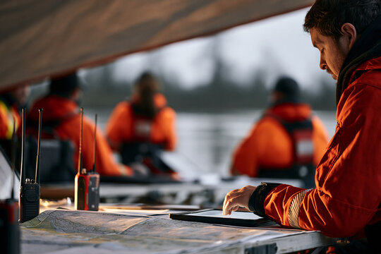 emergency coordination tent at the edge of a flooded district, maps and radios on folding tables, responders in hi-vis seen from behind, overcast grey light, standing water beyond, cinematic photoreal