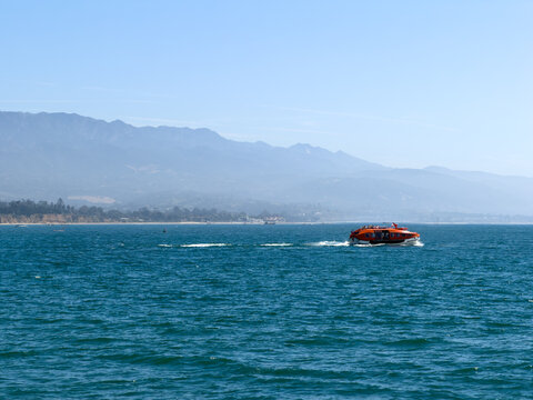 SANTA BARBARA, CA, APR 8, 2026: An orange tender shuttle boat speeds across the water toward a cruise ship, with hazy mountains rising in the background