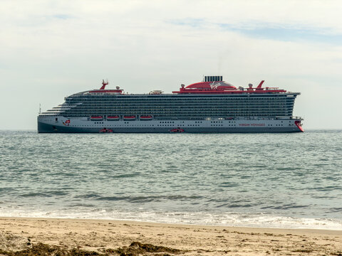 Santa Barbara, CA, APR 9, 2026: Virgin Voyages cruise ship rests offshore near a sandy beach, its distinctive red funnel visible under an overcast sky