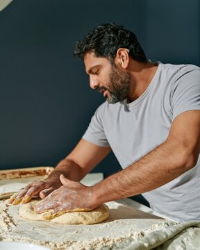 Hands of a South Asian man kneading dough on a floured board, featuring powdery textures and soft window light for home baking and inclusive kitchen campaigns