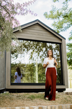 Joyful woman stands in front of modern cabin with big glass window