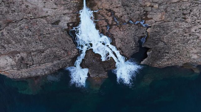 Industrial wastewater cascades from a reverse osmosis desalination plant outlet down rugged limestone cliffs into the sea, captured by drone above Ghar Lapsi, Malta.