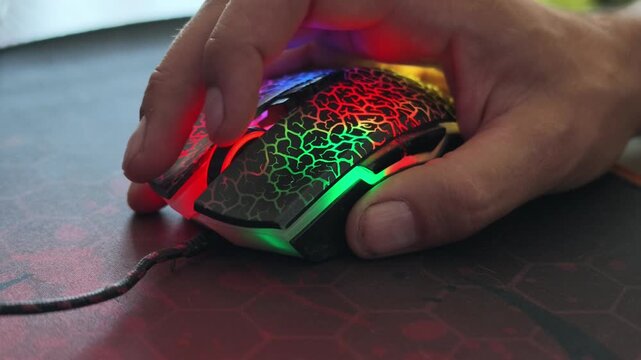 A hand grips a cracked, rainbow-lit gaming mouse on a dark hexagonal pad, repeatedly pressing buttons with subtle RGB pulses.