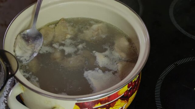 A hand uses a spoon to skim foam from a simmering pot of meat broth, repeatedly lifting impurities while the liquid bubbles gently on the stovetop.