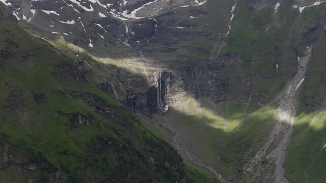 Narrow waterfalls drop down a shaded alpine cirque as shifting sunlight moves across the steep rock face and grassy slopes.
