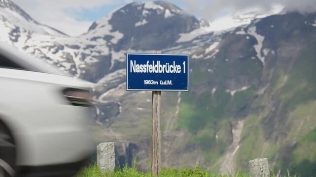 Nassfeldbr&uuml;cke 1 altitude sign stands on a grassy verge with snow-streaked mountains rising behind it in soft daylight.