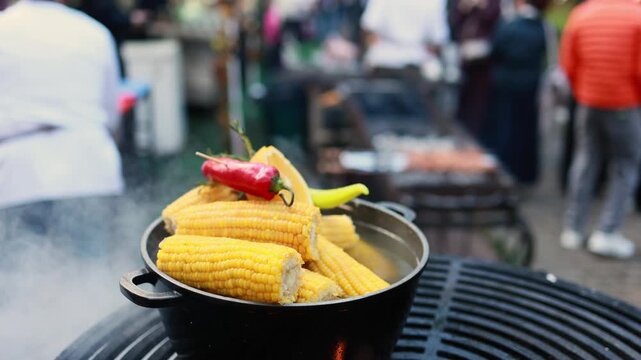 Boiled corn cobs in cast iron pot with chili peppers at street food festival. Outdoor cooking on grill with smoke and blurred people. Street food market concept.
