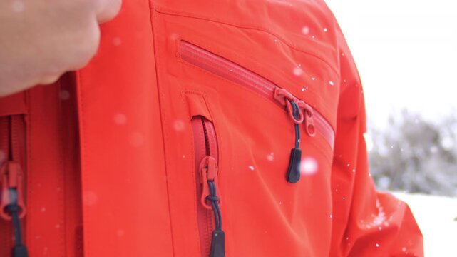 Person pulls zipper tab to open a red winter jacket while standing outdoors in the snow