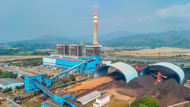Aerial view of a large coal-fired power plant featuring a tall chimney, blue industrial buildings, conveyor belts, and large coal storage domes