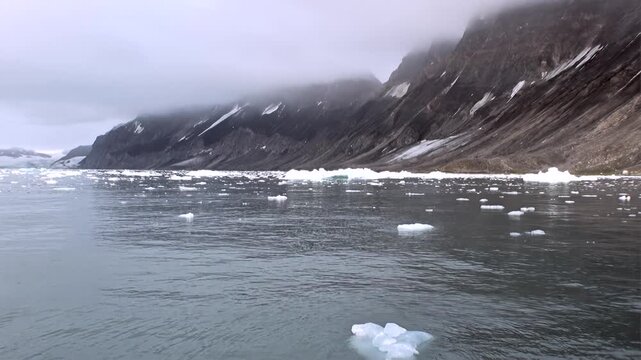 Ice Floes Drift Below Foggy Mountain Slopes in Svalbard Arctic. Broken ice moves across cold coastal water beside steep dark mountains partly covered by low cloud and lingering snow.