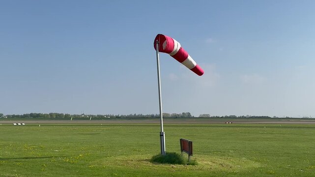 A red and white windsock stands beside a grassy airport runway under a clear blue sky, indicating wind direction and supporting safe aircraft operations.