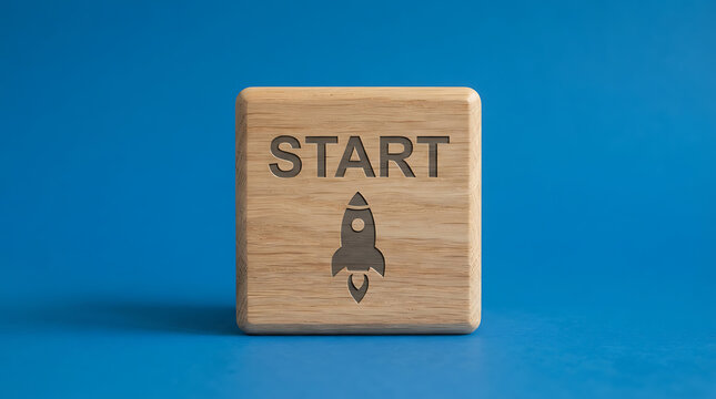 A square wooden block with the word start and a small rocket ship icon engraved on the front, placed on a solid blue background, representing the beginning of a project, journey, or new venture