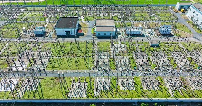 Aerial view of an electrical substation with transformers, switchgear, utility buildings, green grass, concrete paths, and long shadows in bright sunlight.