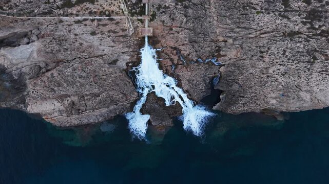 Industrial wastewater cascades from a reverse osmosis desalination plant outlet down rugged limestone cliffs into the sea, captured by drone above Ghar Lapsi, Malta.