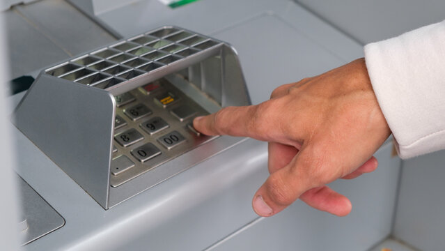 Hand presses keypad on ATM machine. User enters PIN with focused intent. Silver machine shows modern banking access.