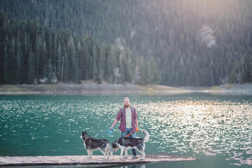 Bearded man in a plaid shirt walks two Siberian husky dogs along the shore of a turquoise mountain lake framed by dark autumn spruce forest, hiking and travelling with pets concept © iwavephoto