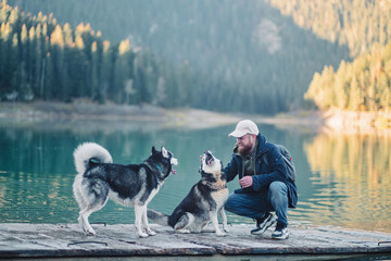Bearded man kneels face to face with two Siberian husky dogs on a pier by a mountain lake in autumn, a close warm frame about affection between a dog owner and his animals © iwavephoto