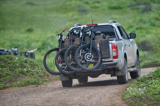 A pickup truck rolls down a dirt trail through lush green hills, its bed packed with five downhill mountain bikes lined up wheel to wheel, race plates and knobby tires facing the camera. 