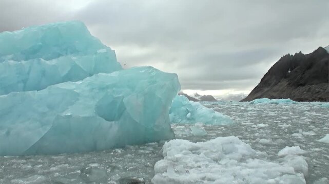 Massive Blue Iceberg Rises Among Brash Ice in Svalbard Arctic. A vivid turquoise iceberg towers beside a field of smaller ice fragments, creating a dramatic polar seascape under gray cloud.