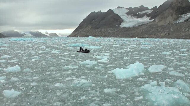 Small Expedition Boat Crosses Ice-Filled Svalbard Arctic Water. A dark inflatable boat carrying people moves among scattered ice floes beneath steep snowy mountains in the polar summer.