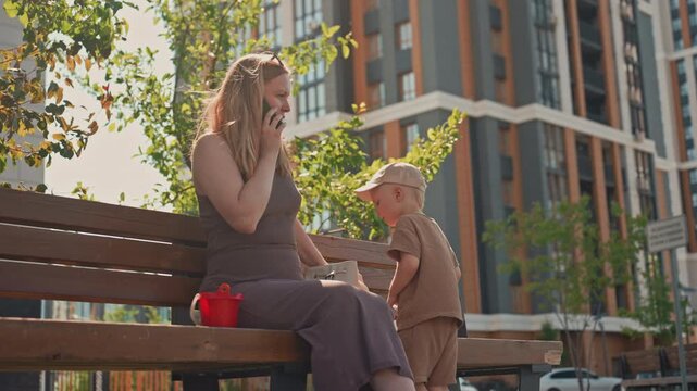 Woman sits on wooden bench in urban park talking with her cell phone while young boy stands nearby holding something. scene features green trees modern high rise buildings and bright