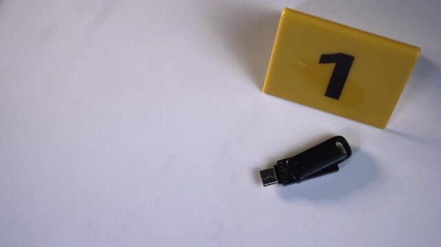 Black USB flash drive next to yellow evidence marker 1 on clean white marble surface for digital forensics investigation with copy space, handheld top view POV shot