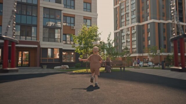 Young boy runs towards woman sitting on bench in outdoor playground holding something small. setting features modern apartment buildings with large windows and balconies under bright