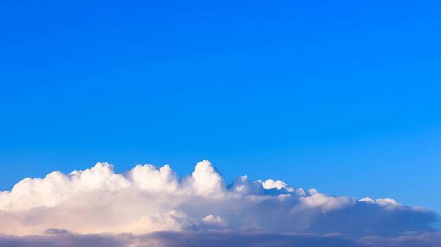 White cumulus clouds slowly drift across a vivid blue sky above a distant cloud bank, forming a bright panoramic weather scene with open atmospheric space and soft natural light