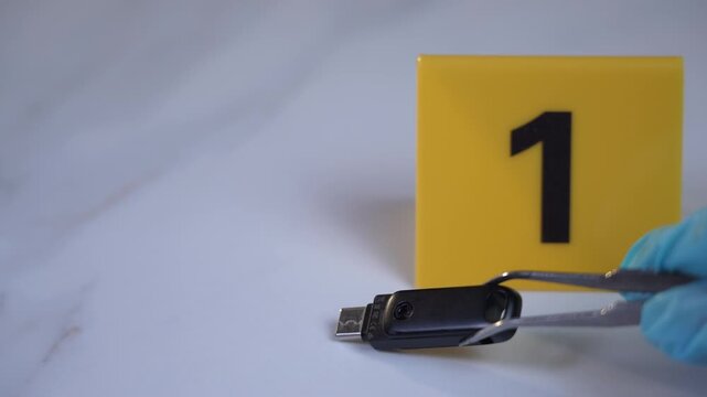 USB drive picked up by gloved hand using tweezers at yellow evidence marker 1 on white marble surface for digital forensics cyber crime investigation, 45 degree close up shot