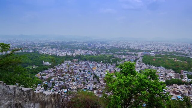 A stone parapet and hillside vegetation frame a wide view over Udaipur's buildings, roads and green urban spaces.