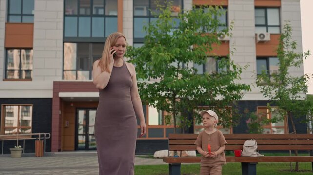 Woman walks while talking on her phone accompanied by young boy holding orange object. scene takes place outdoors in front of modern building with greenery and benches visible in background