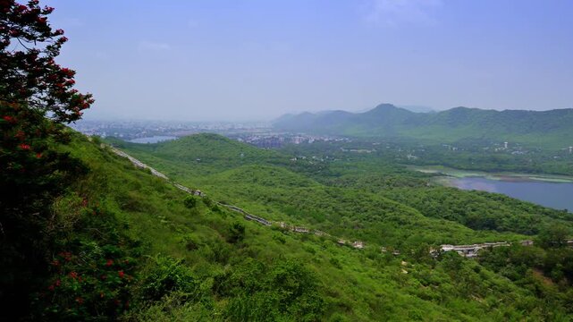 Green ridges and a winding hillside wall extend toward Udaipur, with a sliver of lake visible beside the wooded slopes.