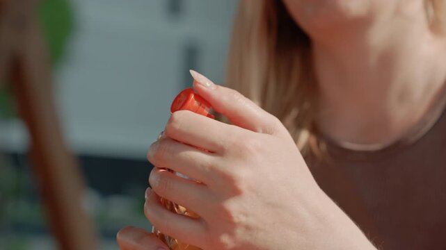 Woman unscrews cap of plastic bottle with orange liquid revealing it to be juice. scene is set outdoors in daylight focusing on her hands and upper body as woman interacts with drink