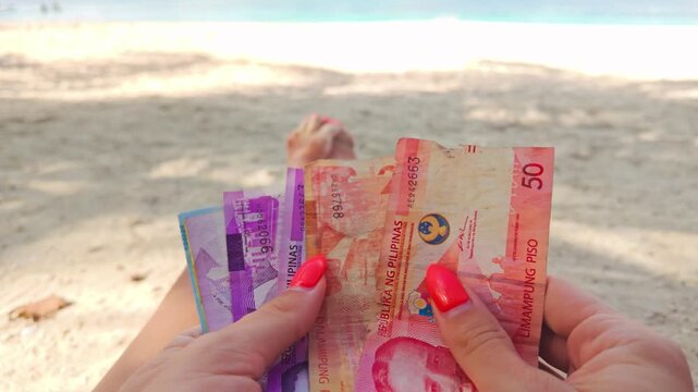 Woman counts colorful Philippine peso notes by hand on a sunny beach shore