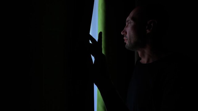 A man in a dimly lit room pulls back a curtain to reveal a sliver of bright green and blue light, then lets it fall back into darkness.