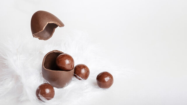 Cracked chocolate easter egg with chocolate small round candies and flying eggshell in white feather nest, levitation