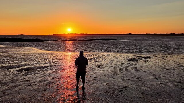 A person walks through shallow tidal water at sunset with brilliant reflections across the Atlantic shore