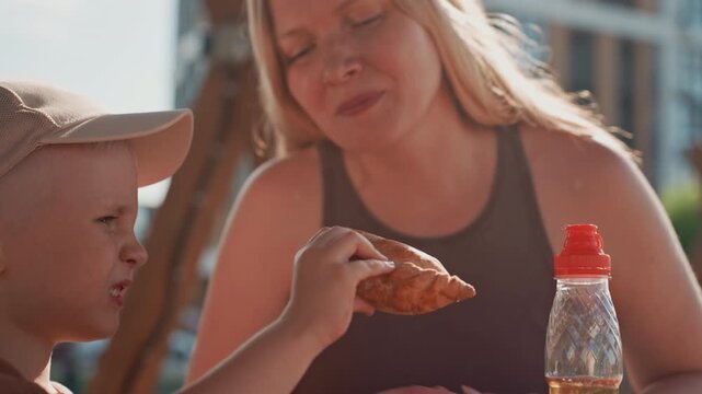 Young boy and woman share food outdoors with child holding bread while adult offers it. plastic bottle is visible on table in soft daylight outdoor lighting