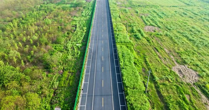 Aerial view of a straight asphalt road crossing lush green fields and woodland, with yellow centerlines, white edge markings, utility poles, and bright sunlight.