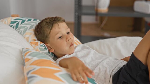 Boy on bed kicking leg and leaning back, reaching toward shelf amid patterned pillows in bedroom; playful childhood moment.