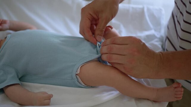 Careful father's hands buttoning the snap fasteners on his baby boy's blue bodysuit. Loving parent getting his newborn son dressed on a changing table in a brightly lit room