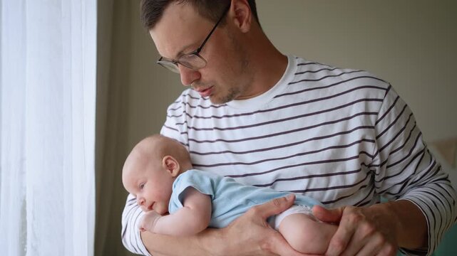 Young, attentive father with glasses gently rocking his sleepy newborn baby, providing comfort and care. The man cradles the infant in his arms near a window in a softly lit room at home