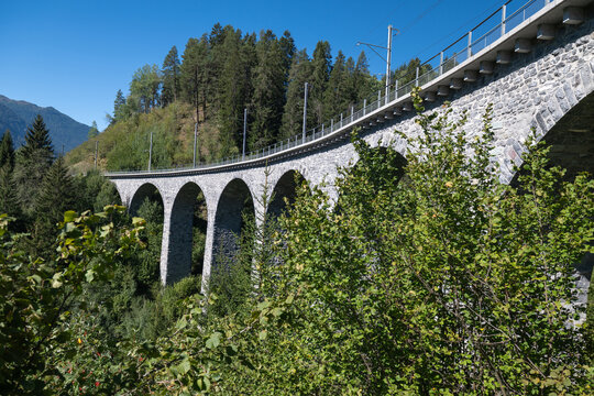 Historic stone railway viaduct curving through lush alpine forest near Filisur, Switzerland, beneath a clear blue sky.