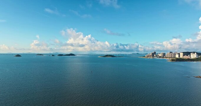 Aerial view of blue coastal water with scattered islands, a distant bridge, and a high-rise waterfront city under bright sunlight and clouds.