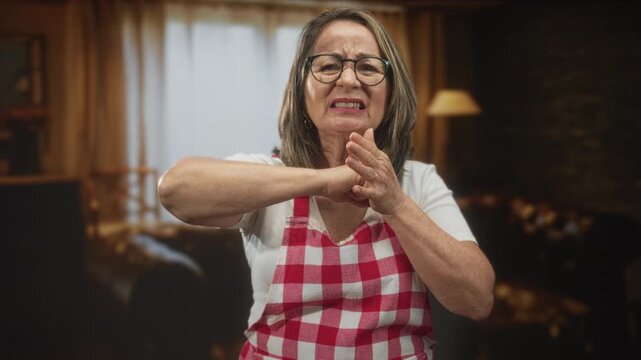 Hispanic woman rubbing wrist and wincing while wearing red check apron in living room; pain aging resilience.