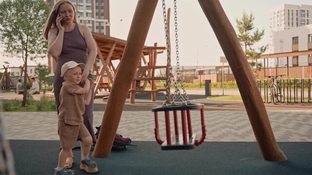 Woman talks on her phone while holding hands with young boy at outdoor playground. child interacts near swings and play structures wearing casual clothing including cap. Other people walk